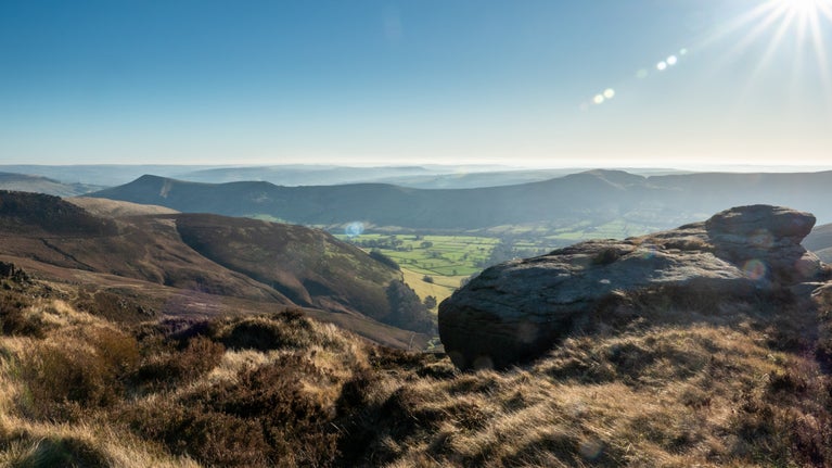 A view across the gritstone tors at Kinder Scout on a sunny day, with the moor visible in the foreground looking out over several tors.